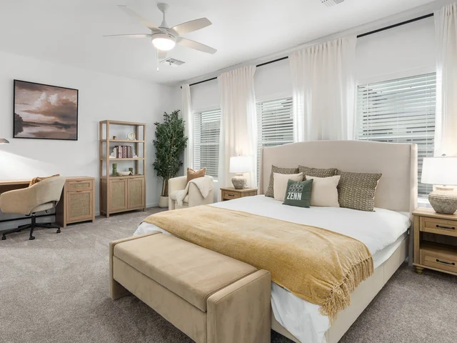 Sunlit bedroom with a neutral upholstered bed, wood furniture, and a desk